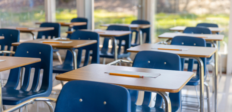 A row of empty classroom desks with a pencil and paper on top of the desk.